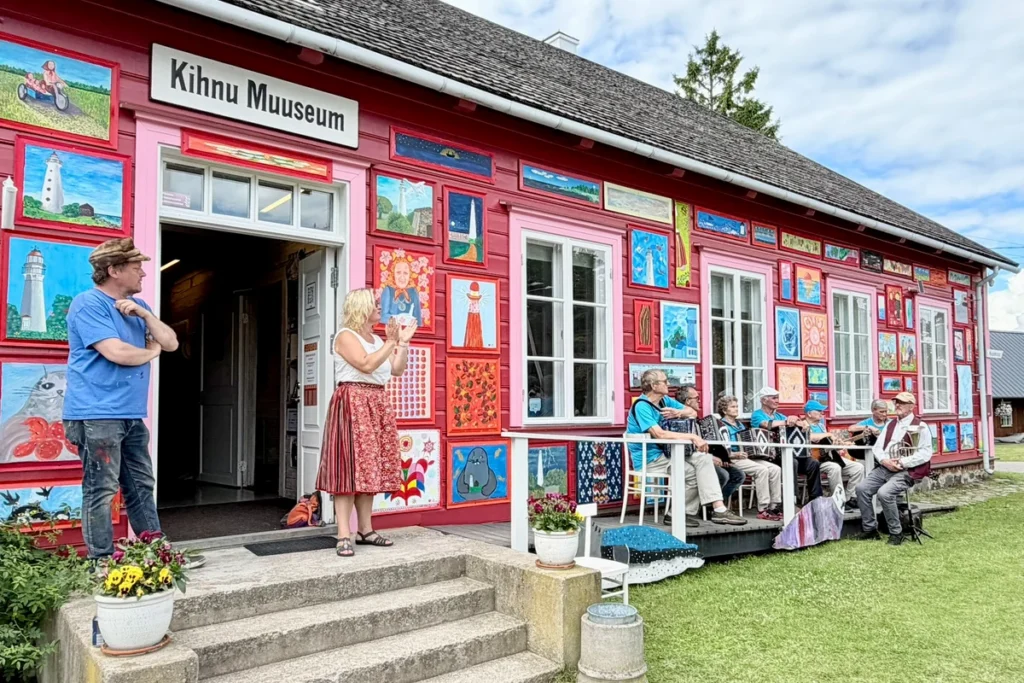 Kihnu Muuseumi roosakaspunane välissein on kaetud värvikirevate maalidega, toimub „Tere TuleMast!” näituse avamine, esinevad pillimehed.