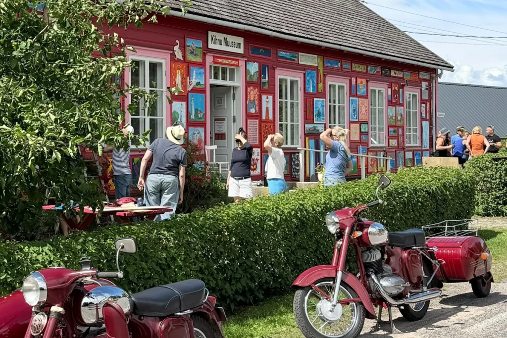 Kihnu Muuseumi roosakaspunasele välisseinale paigaldatud värvikirevad maalid näituse „Tere TuleMast!” ülespaneku ajal. Esiplaanil kaks punast külgkorviga mootorratast.