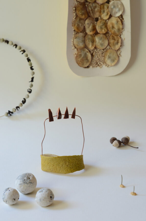 At the centre of the photograph is a ring made from pomelo peel, topped with a wire frame bearing triangular pieces cut from an avocado pit. Around it are beads made from flour, along with other forms created from seeds and plant-based materials.