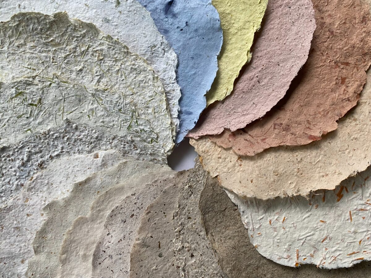 Handmade paper sheets in varied colors overlap in close-up, with plant matter visible in the paper.