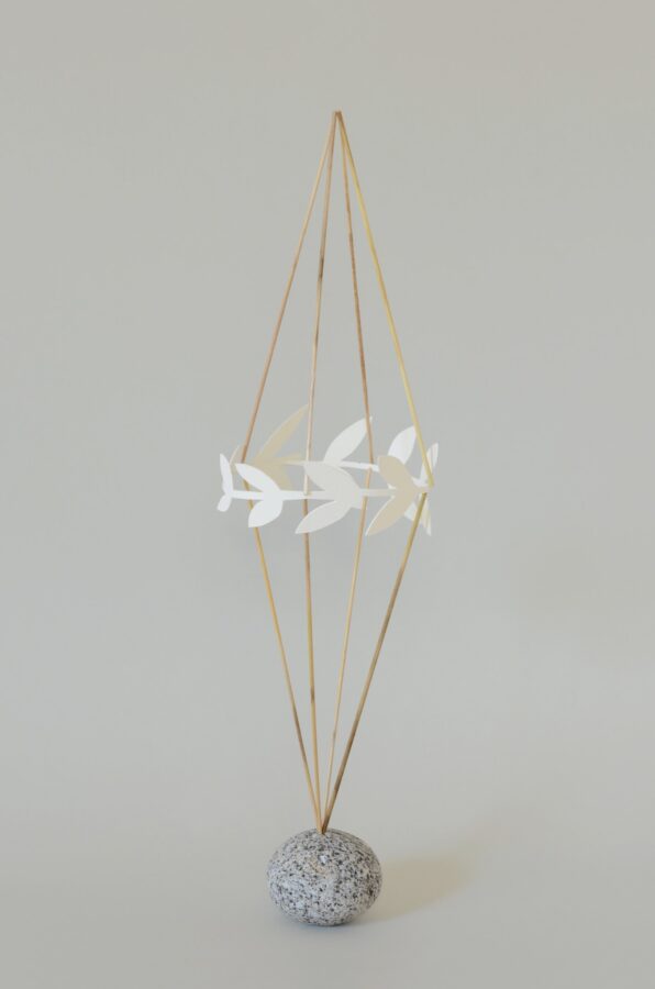 An elongated form made from wood small-reed (Calamagrostis epigejos) stems, with leaf shapes cut from natural-white watercolour paper at its centre. The form rests on a rounded grey granite stone.